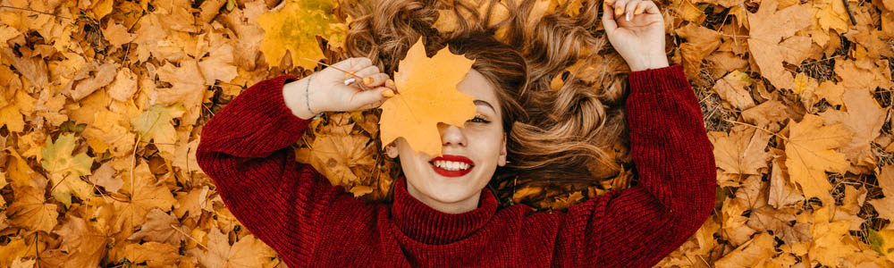 Young, beautiful woman with red lipstick laying on her back in a pile of autumn leaves, holding one leaf up in front of her face